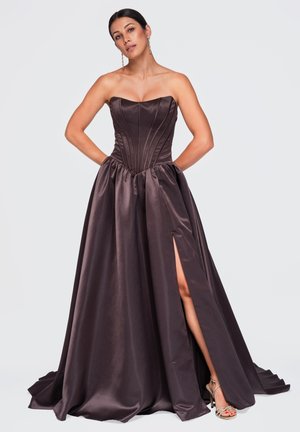 Femme portant une robe bustier en satin brun foncé avec un corsage ajusté et une fente haute sur la cuisse, posant les mains dans le dos, portant des boucles d'oreilles pendantes.