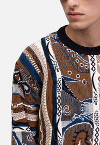 Strickpullover mit geometrischen Mustern in Braun, Blau, Schwarz und Weiß. Gerippter schwarzer Kragen; strukturierte Gestaltung mit unterschiedlichen Sticktechniken.