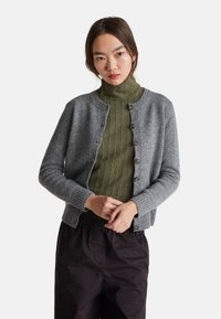 Šedý vlněný cardigan s žebrovanými manžetami a lemem, s šesti předními knoflíky a kulatým výstřihem, nošený přes tmavě zelený strukturovaný svetr.