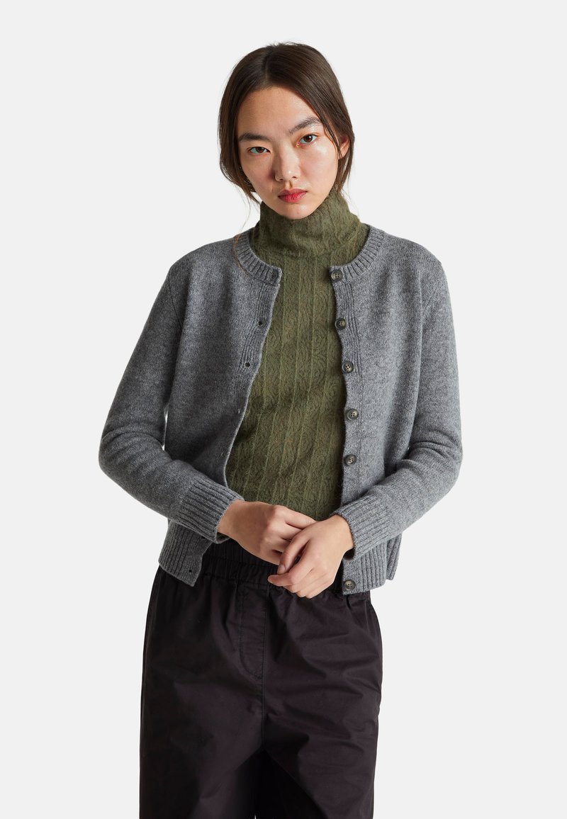 Šedý vlněný cardigan s žebrovanými manžetami a lemem, s šesti předními knoflíky a kulatým výstřihem, nošený přes tmavě zelený strukturovaný svetr.