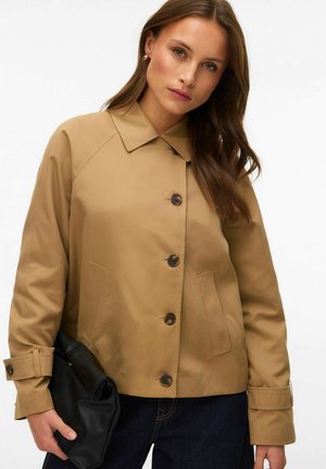 Femme portant une veste beige boutonnée tenant une pochette noire pliée, cheveux longs et bruns, debout devant un fond blanc.
