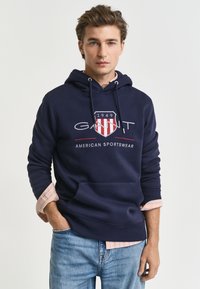 Felpa navy con cappuccio caratterizzata da un logo frontale con accenti rossi e bianchi, cappuccio con cordino e tasca a marsupio, indossata sopra una camicia a quadri chiari.