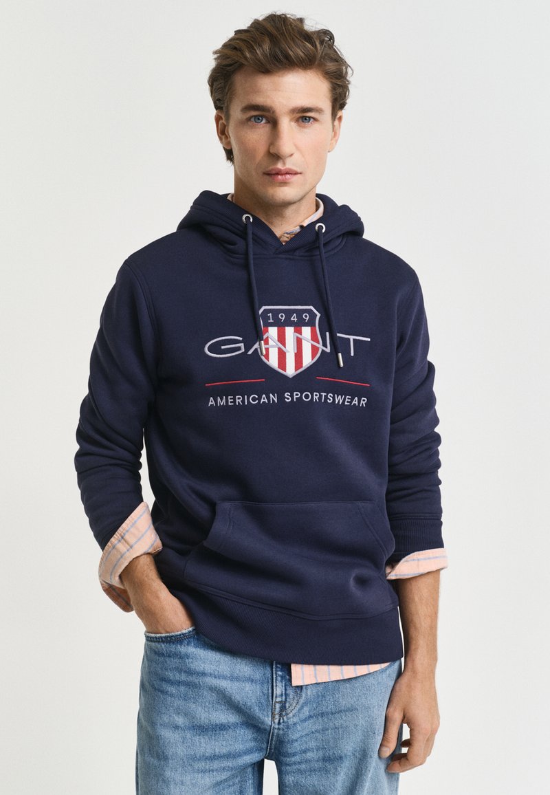 Navy blue hoodie featuring a front logo with red and white accents, drawstring hood, and a kangaroo pocket, layered over a light plaid shirt.