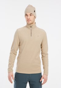 Pullover in fleece beige con collo alto e chiusura a zip. Berretto a coste beige indossato da un modello maschile. Tessuto morbido, design semplice, minimalismo del brand.