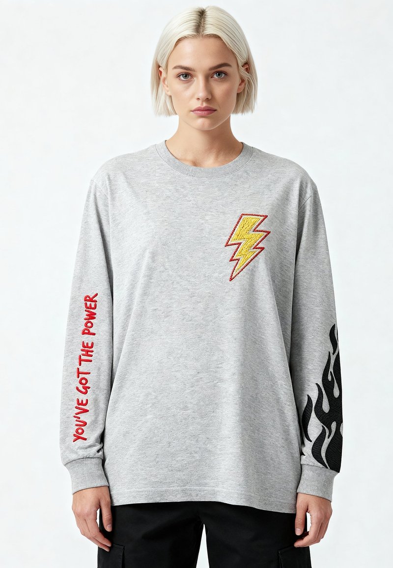 Junge Person mit kurzen blonden Haaren, trägt ein graues Langarmshirt mit einem gelben Blitz, rotem Text am linken Arm und schwarzen Flammen am rechten Arm.