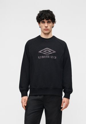 Ung mann med krøllete hår iført en svart Umbro-genser og svarte bukser, stående foran en enkel lys grå bakgrunn.