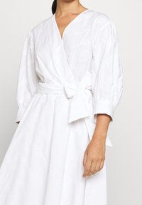 Robe enveloppante blanche en coton avec des manches bouffantes, un design à œillets texturé et une ceinture à nouer pour un ajustement réglable.