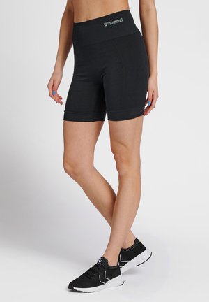 Vrouw draagt zwarte nauwsluitende Hummel-shorts en zwarte sportschoenen, staand tegen een effen lichte achtergrond.