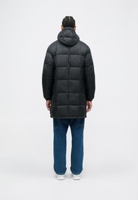 Cappotto lungo imbottito nero con cappuccio, caratterizzato da sezioni trapuntate e una texture liscia, abbinato a pantaloni in denim azzurri larghi e scarpe bianche.