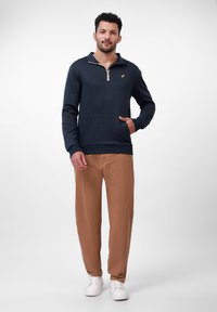 Mørkeblå teksturert genser med kvart-glidelås krage, gyldent logo detaljer, og ribbestrikkede mansjetter, parret med brune bukser og hvite sneakers.