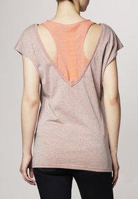T-shirt à manches courtes avec une coupe ample, de couleur rose clair, dos en V avec une découpe, et un débardeur orange visible en dessous.