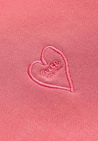 Różowy materiał z haftowanym serduszkiem na środku, z napisem "Cute" wewnątrz. Gładka tekstura, średnia waga, styl casual.