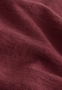 Gros plan d'un tissu doux et texturé de couleur bordeaux profond avec un motif de tissage visible et des plis doux créant des ombres et de la profondeur.