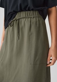 Close-up of waist wearing olive green skirt with elastic drawstring and large front pockets, paired with a tucked-in black top.