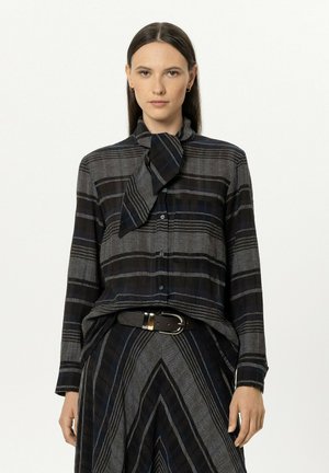Woman wearing a black and gray striped blouse with a tied scarf neckline and matching skirt with a black belt, standing against a white background.