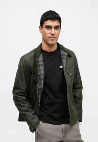 Jachetă bomber verde măsliniu cu guler de catifea, închidere cu fermoar și căptușeală interioară în carouri, purtată peste un hanorac negru. Textură netedă.