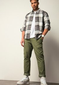 Gestreepte knoopshirt in grijs en wit over een wit T-shirt, gecombineerd met olijfgroene cargobroeken en witte sneakers met grijze accenten.
