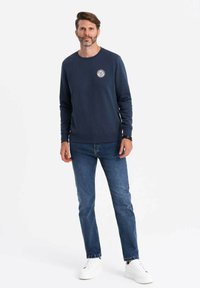 Sudadera de algodón azul marino con cuello redondo, puños y dobladillo acanalados, que presenta un parche de logo circular en el pecho. Combinada con jeans azules y zapatillas blancas.