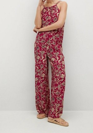 Kvinde iført ærmeløs bordeaux jumpsuit med beige blomsterprint og beige flade sandaler med remme står op ad en lys væg.