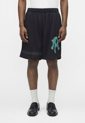 Férfi, aki fekete, rugalmas derékpánttal ellátott rövidnadrágot visel, zöld logóval a bal lábszáron, fehér zoknival és fekete mokaszin cipővel.