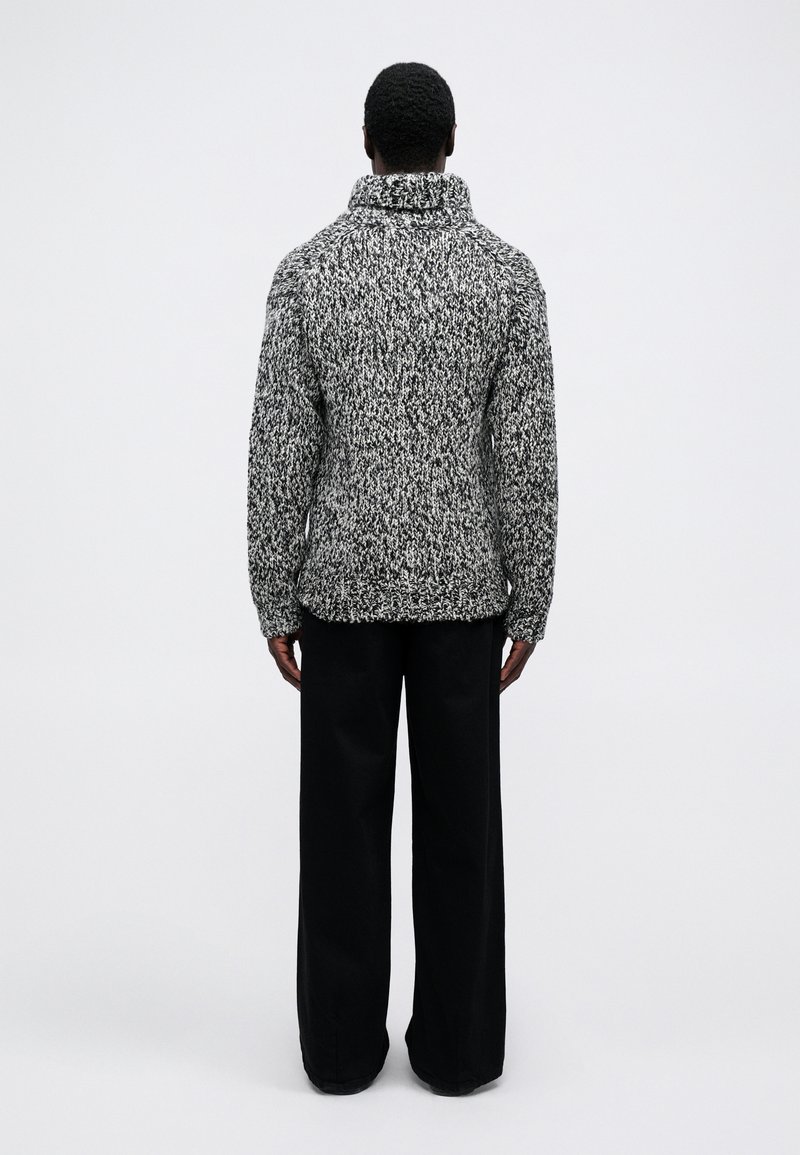 Maglione bianco e nero testurizzato con colletto alto, vestibilità ampia e polsini a costine, abbinato a pantaloni neri a gamba larga.