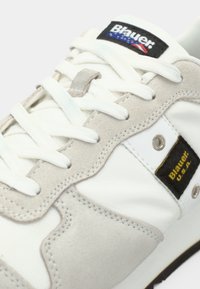 Baskets blanches et grises avec lacets blancs, avec des étiquettes noires indiquant Blauer USA sur la languette et le panneau latéral.