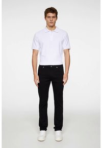 Biała koszulka polo z krótkimi rękawami, zestawiona z czarnymi obcisłymi jeansami i białymi trampkami, stojąca prosto na jednolitym tle.