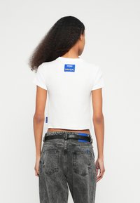 Λευκό κοντό μπλουζάκι με κοντά μανίκια, με μπλέ λογότυπο στο επάνω μέρος της πλάτης. Συνδυασμένο με γκρι τζιν παντελόνι, εμφανείς ραφές και δύο πίσω τσέπες.