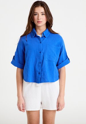 Jonge vrouw staand, draagt een felblauw overhemd met knopen en opgerolde mouwen en witte gestructureerde shorts tegen een effen witte achtergrond.