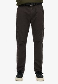 Un pantalon cargo en tissu marron foncé, doté de plusieurs poches latérales, d'une texture lisse, d'une coupe fuselée, et associé à des baskets noires.