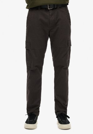 Un pantalon cargo en tissu marron foncé, doté de plusieurs poches latérales, d'une texture lisse, d'une coupe fuselée, et associé à des baskets noires.