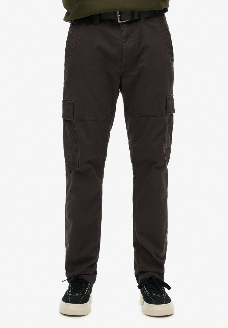 Un pantalon cargo en tissu marron foncé, doté de plusieurs poches latérales, d'une texture lisse, d'une coupe fuselée, et associé à des baskets noires.