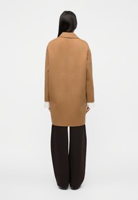 Καμηλό παλτό από μαλλί με ευθύγραμμη σιλουέτα, γιακά με άνοιγμα σε V και μακριά μανίκια, φορεμένο πάνω από σκούρο παντελόνι με φαρδιά μπάτζα.