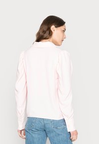 Camisa de manga longa rosa claro com ombros bufantes, punhos com botões e colarinho. Tecido texturizado e design nas costas reto. Usada com jeans azuis.