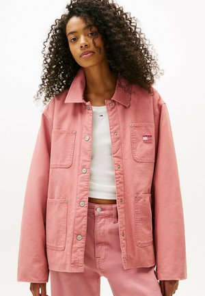 Veste en denim rose avec deux poches avant, boutons-pression en métal argenté et col. Portée par-dessus un haut blanc, avec un pantalon rose assorti.
