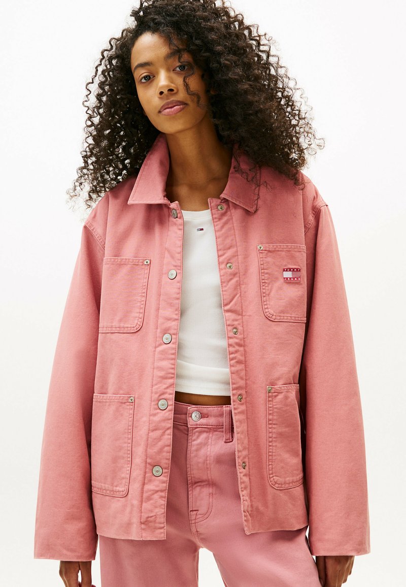 Giacca di denim rosa con due tasche frontali, bottoni a pressione argento e colletto. Indossata sopra una maglietta bianca, con pantaloni rosa abbinati.