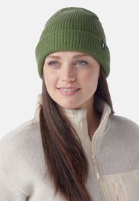 Grüne Strickmütze mit geripptem Muster und einem kleinen Logoschild. Das Modell trägt eine leicht beige Fleecejacke mit Reißverschluss.