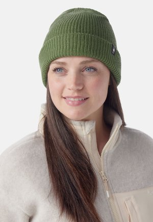 Bonnet en tricot vert avec une texture côtelée et une petite étiquette logo. Le modèle porte une veste en polaire beige clair avec fermeture éclair.