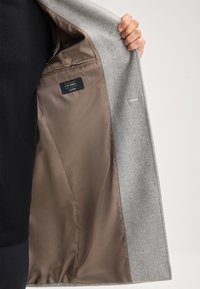 Hand die een open grijs wollen jas toont met een gladde bruine binnenvoering en een ritszakje met merklabel.