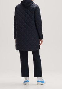 Tummansininen tikkivuorattu pitkä takki hupulla. Siinä on suora leikkaus, kulmikkaat saumat ja se on yhdistetty tummiin housuihin sekä valkoisiin lenkkitossuihin, joissa on sinisiä yksityiskohtia.