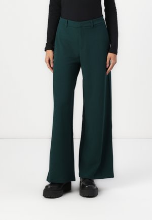 Personne portant un pantalon large vert foncé, des chaussures compensées noires et un haut noir ajusté à manches longues, debout devant un fond blanc.