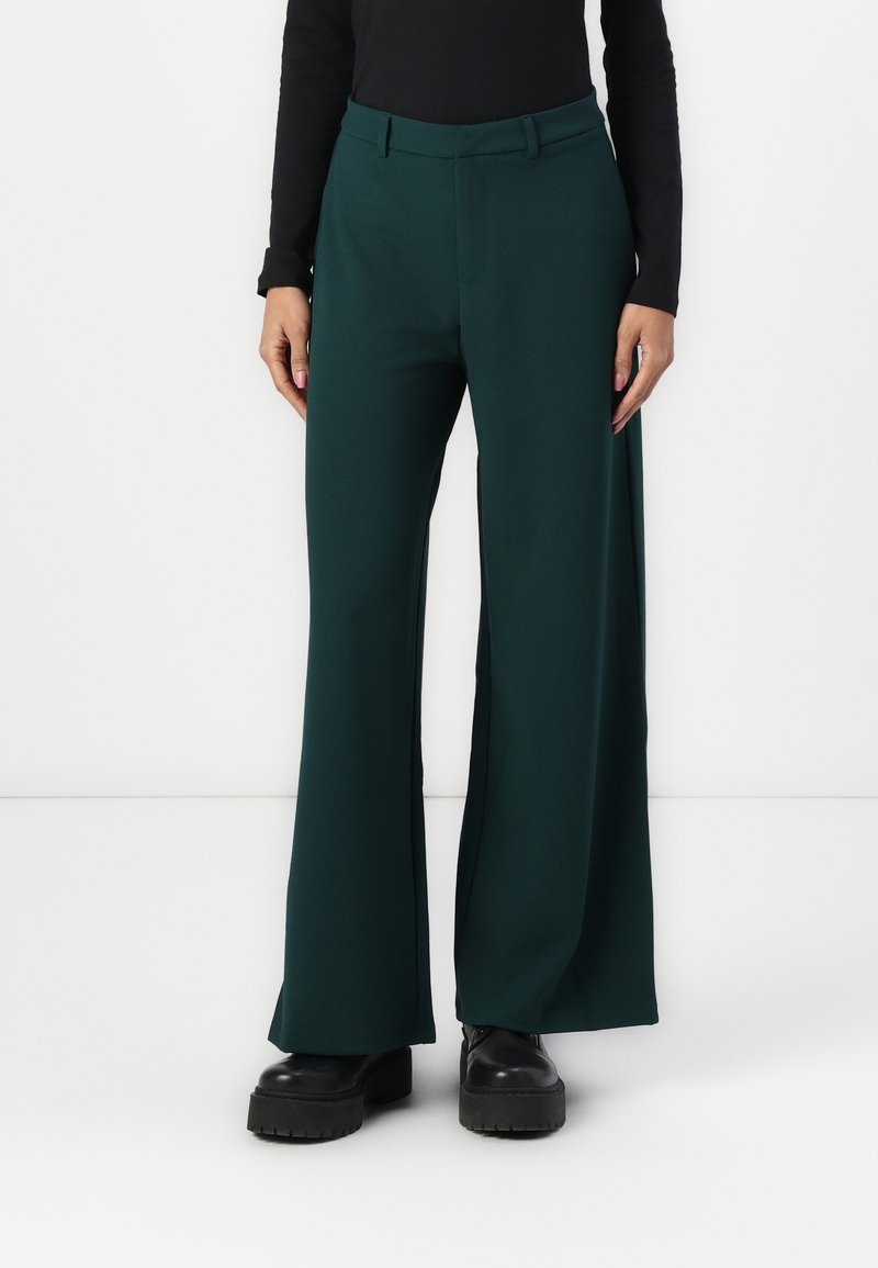 Personne portant un pantalon large vert foncé, des chaussures compensées noires et un haut noir ajusté à manches longues, debout devant un fond blanc.