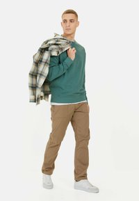Green sweatshirt with a crew neck, beige cargo pants, and white sneakers. A plaid shirt is draped over the shoulder. Clean, casual look.
