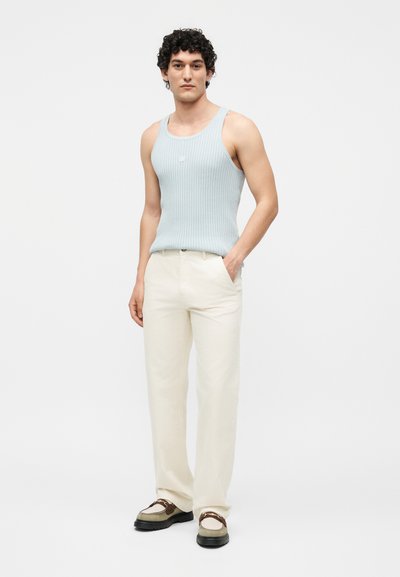 Νέος άνδρας που στέκεται με το χέρι στην τσέπη, φοράει ανοιχτό μπλε αμάνικο ριμπ τοπ, κρεμ παντελόνια και καφέ-λευκά μοκασίνια σε λευκό φόντο.
