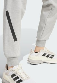 I pantaloni grigi di felpa presentano una striscia verticale nera sul lato. Le sneakers bianche con accenti grigi e neri completano l'outfit.