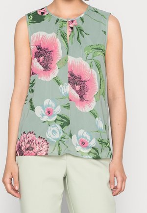 Mouwloze groene blouse met grote roze, witte en groene bloemenprint, gedragen met lichtgekleurde broek.