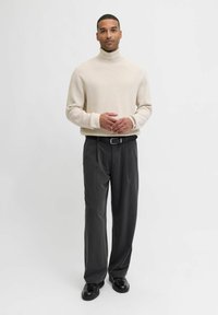 Bež turtleneck-sviiter, vabalt lõikega, pehme tekstuur, koos tumedate hallide pükstega, sirge lõige, must vöö, läikivad kingad, ühtlane taust.