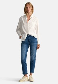 Witte button-up shirt met lange mouwen, borstzak, gecombineerd met medium-wash denim jeans en lichtgekleurde sneakers.