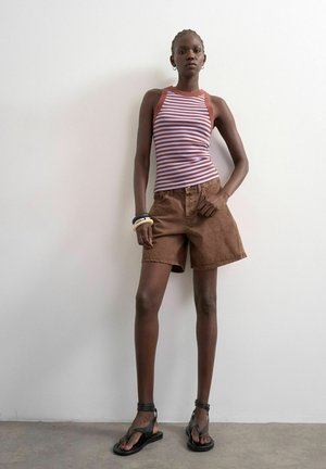 Woman standing against a white wall wearing a sleeveless striped top, brown shorts, black sandals, and stacked bracelets on her wrist.