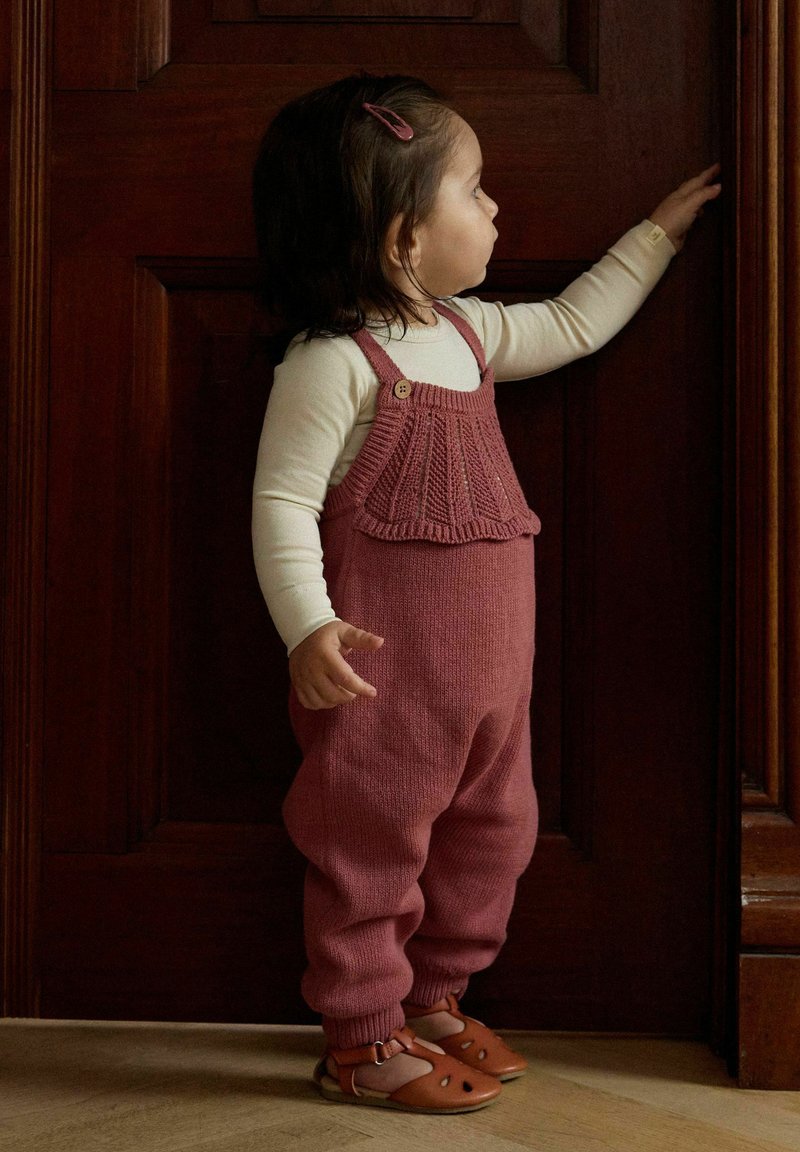 Tout-petit portant une salopette rose et une chemise crème tend la main pour toucher une porte en bois sombre tout en se tenant sur un sol clair.